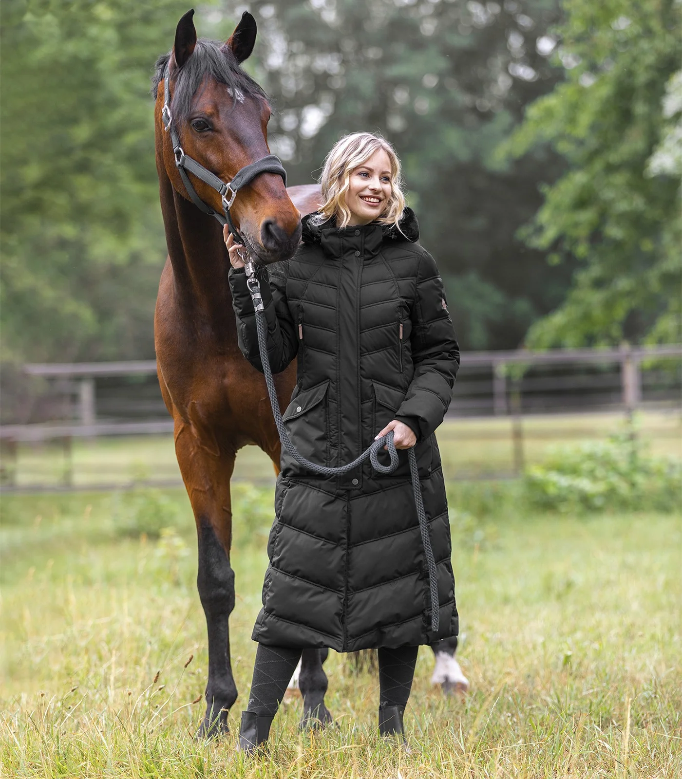 Woman in a black ELT Saphira Winter Ridingcoat holding a horse in a field, showcasing warmth and comfort for riders.
